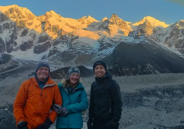 Three happy trekkers posing at Goechala Trek Viewpoint 1 with the snow-covered Kanchenjunga mountain massif at sunrise.